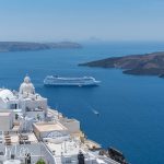 Whitewashed homes spill down the volcanic cliffs toward the deep blue caldera, while cruise ships drift between Santorini and the volcanic islands that mark the center of the ancient eruption. (2879)