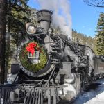 The winter excursion offers a peaceful, intimate view of the landscape, where snow-covered pines, frozen waterfalls, and the rushing Animas River create a striking contrast against the black steam locomotive and its billowing white plume.